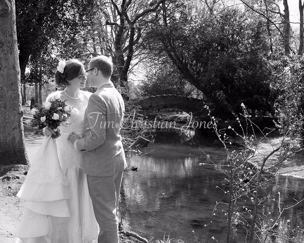 Catherine and Philip stand close, about to kiss, beside the stream at Golden Acre Park near Weetwood Hall, framed by trees and an old stone bridge.