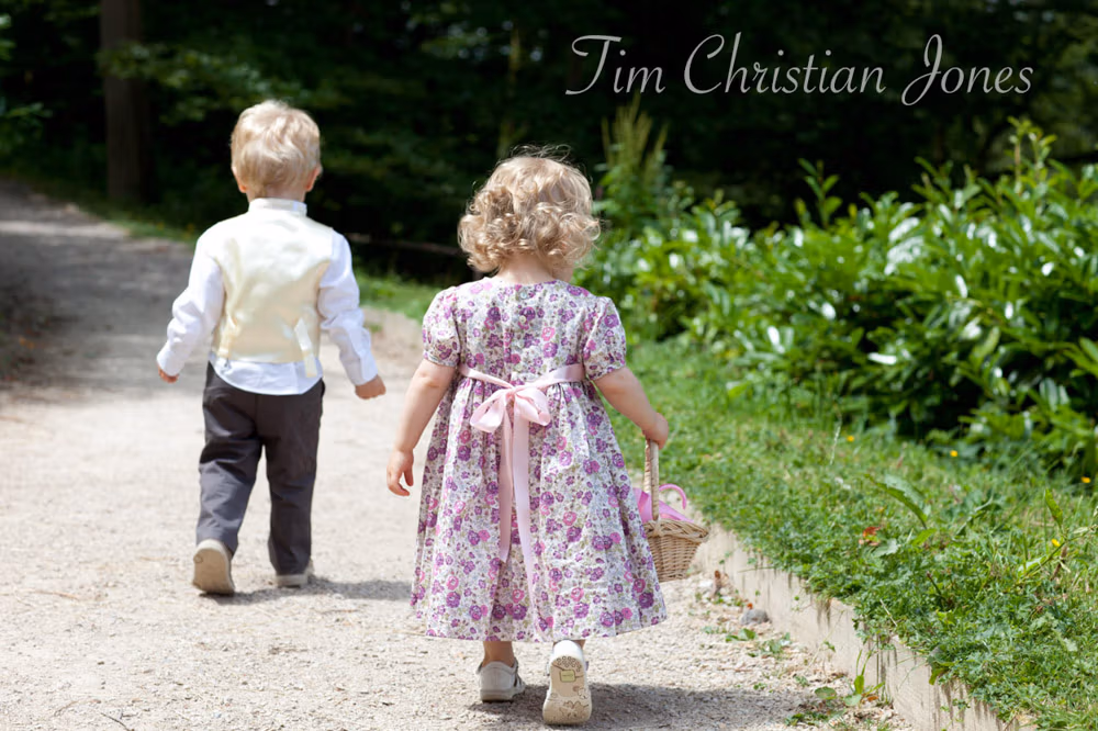 Flower girl walks with the couple’s son along a garden path, basket in hand
