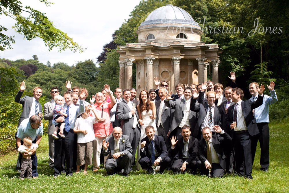 Full wedding party waves in the sun with the Temple of Apollo behind them